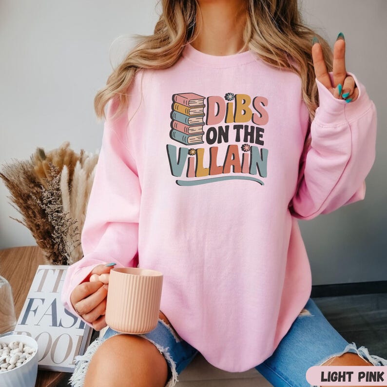 May include: Light pink sweatshirt with the text "Dibs on the Villain" in a colorful, retro font. The design includes a stack of books. A person is wearing the sweatshirt and holding a mug.