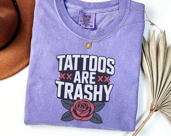 May include: Violet Comfort Colors t-shirt with the text "TATTOOS ARE TRASHY" in white and black, with a red rose graphic. A gold necklace and a brown hat are visible. The shirt is a size small.