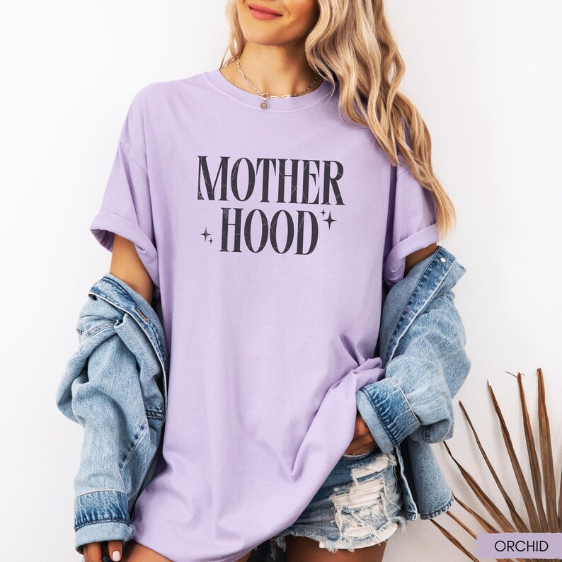 May include: A light purple t-shirt with the text "MOTHER HOOD" in black, with star accents. The shirt is part of a casual outfit, paired with a denim jacket and shorts. The word "ORCHID" is printed in the bottom right corner.