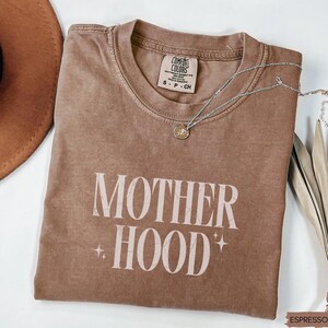 May include: Espresso-colored t-shirt with the word "MOTHERHOOD" in a white, stylized font. The shirt has a crew neck and short sleeves. A silver necklace with a pendant is draped across the shirt.