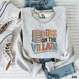 May include: A light gray sweatshirt with the text "Dibs on the Villain" in colorful letters and a stack of books. The sweatshirt is paired with blue jeans, white sneakers, and a gold chain necklace. The word "ASH" is in the bottom right corner.