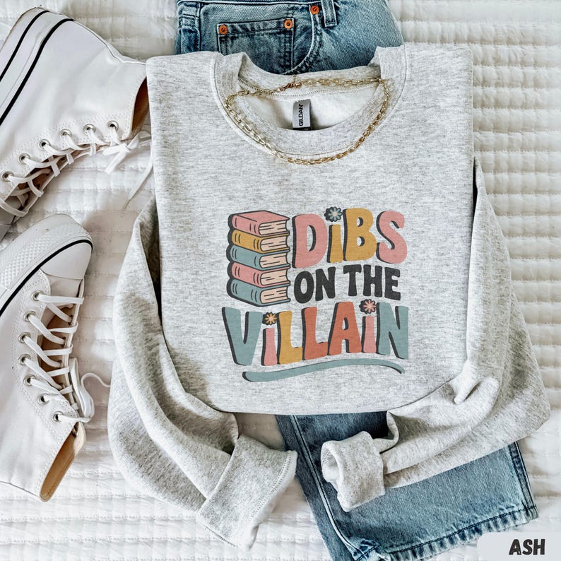 May include: A light gray sweatshirt with the text "Dibs on the Villain" in colorful letters and a stack of books. The sweatshirt is paired with blue jeans, white sneakers, and a gold chain necklace. The word "ASH" is in the bottom right corner.