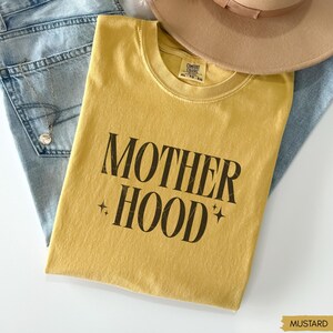 May include: Mustard yellow t-shirt with the text "MOTHER HOOD" in a bold, black font. The shirt is folded and rests on a pair of blue jeans. A tan hat is visible in the background. The shirt is size XL-TG-EG.