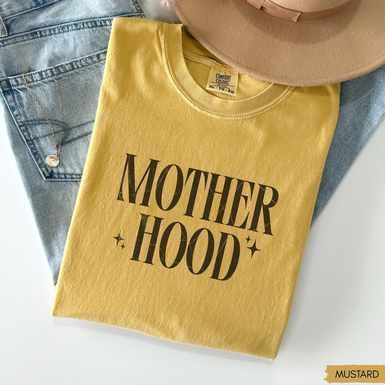 May include: Mustard yellow t-shirt with the text "MOTHER HOOD" in a bold, black font. The shirt is folded and rests on a pair of blue jeans. A tan hat is visible in the background. The shirt is size XL-TG-EG.