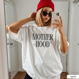May include: White t-shirt with the text "MOTHER HOOD" in a bold, dark gray font. The shirt has short sleeves and a relaxed fit. The model is wearing a red beanie and black sunglasses.