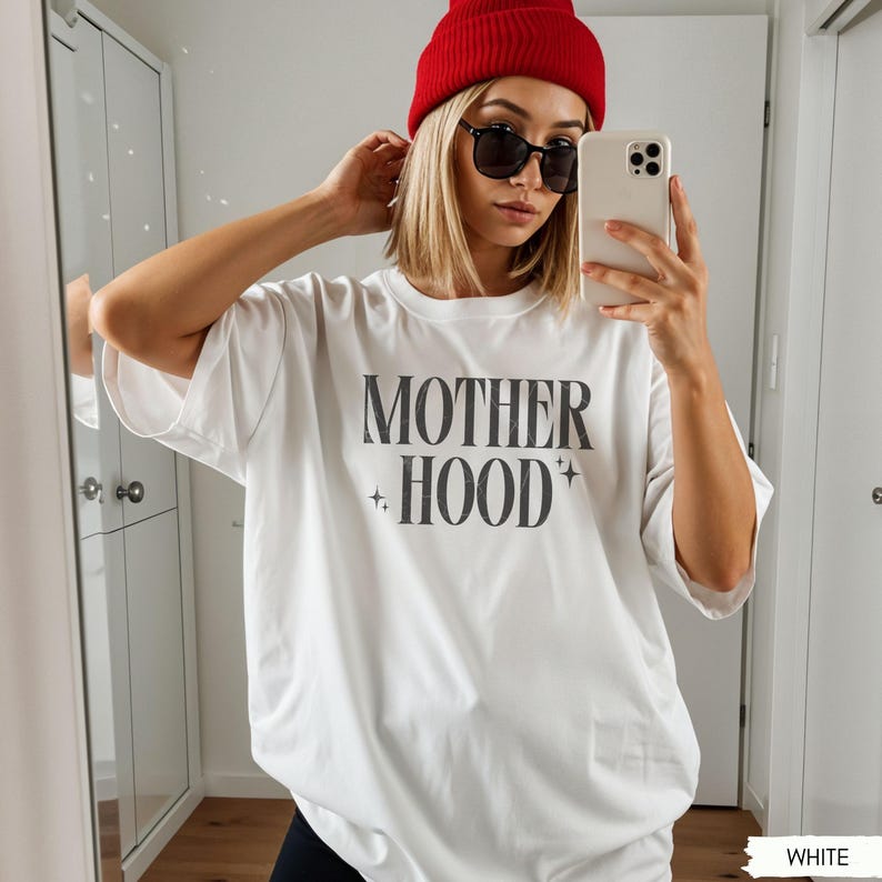 May include: White t-shirt with the text "MOTHER HOOD" in a bold, dark gray font. The shirt has short sleeves and a relaxed fit. The model is wearing a red beanie and black sunglasses.