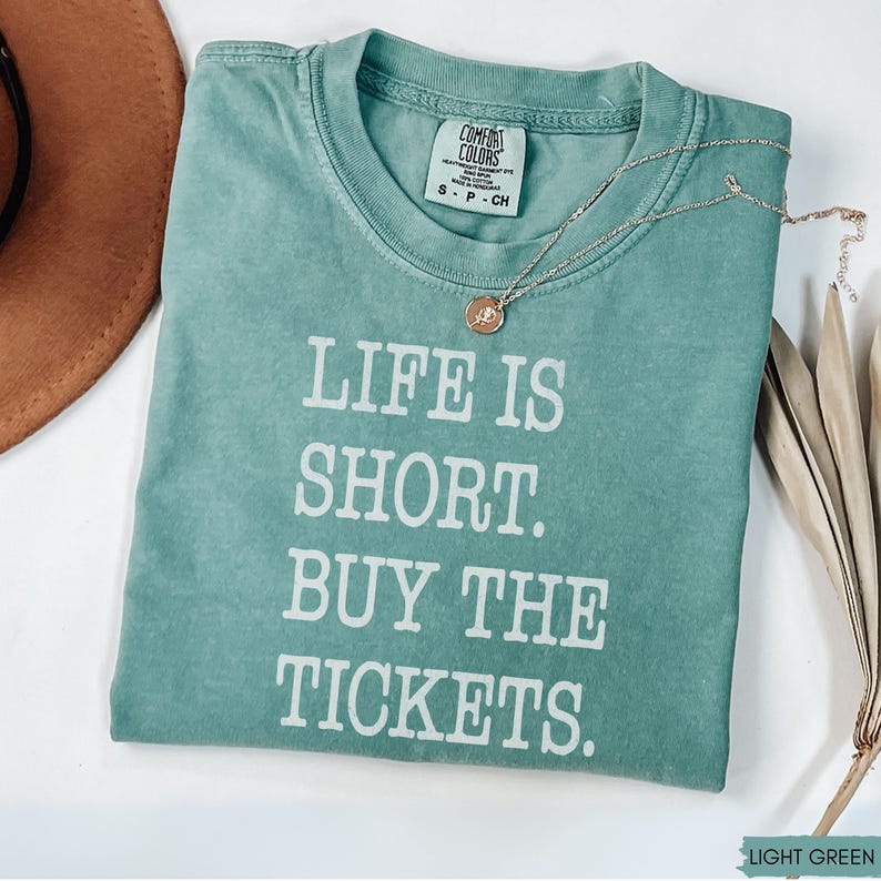 Puede incluir: Una camiseta verde claro con el texto blanco "LIFE IS SHORT. BUY THE TICKETS.". Un collar dorado con un colgante descansa sobre la camiseta doblada. La camiseta tiene una etiqueta Comfort Colors.
