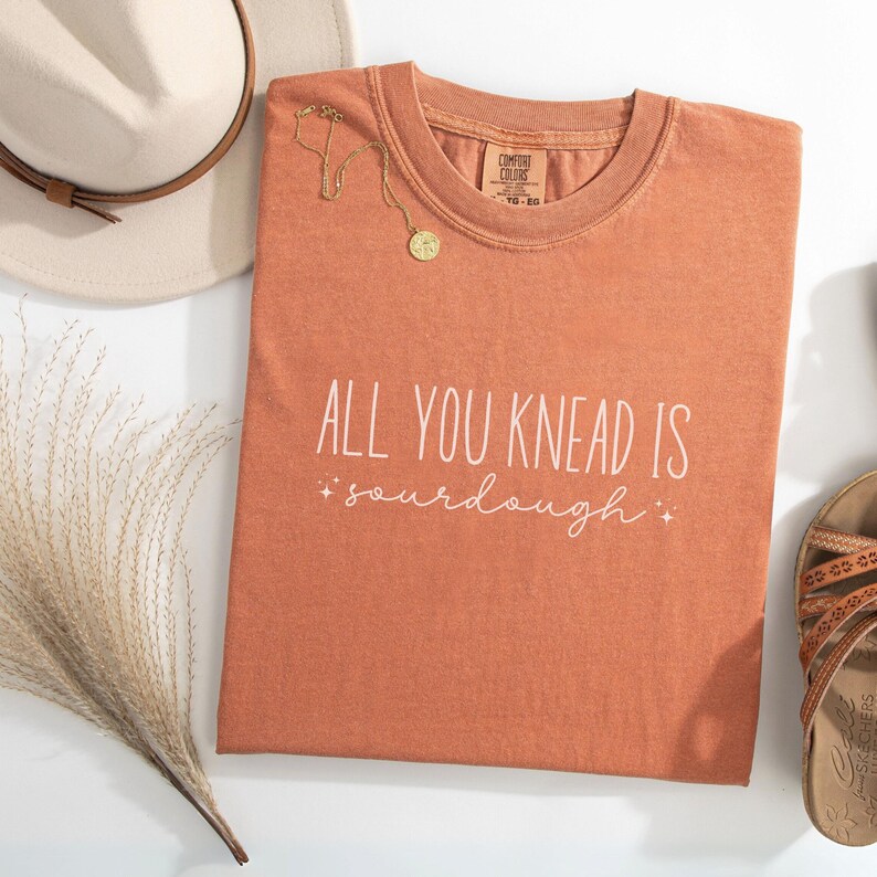 May include: A rust-colored t-shirt with the text "All you knead is sourdough" in white lettering. The shirt has a distressed look and is made of a soft, comfortable fabric.
