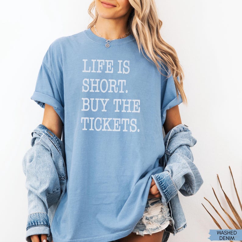 Puede incluir: Una camiseta azul claro con el texto blanco "LIFE IS SHORT. BUY THE TICKETS.". La camiseta tiene las mangas enrolladas y se combina con una chaqueta vaquera. La esquina inferior derecha de la imagen tiene el texto "WASHED DENIM".