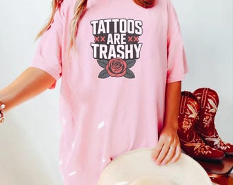 May include: A light pink t-shirt with the text "TATTOOS ARE TRASHY" in a bold, black font, with a rose graphic. The shirt is a casual style, with short sleeves.