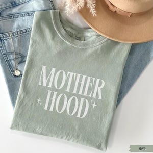 May include: A sage green t-shirt with the word "MOTHERHOOD" printed in white, with star accents. The shirt is laid on top of blue jeans, with a gold necklace and a tan hat visible in the background.