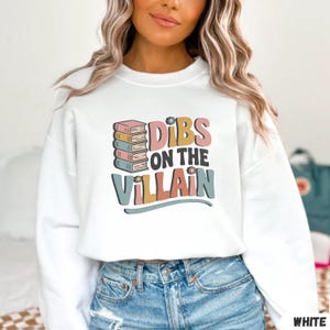May include: White sweatshirt with the text "Dibs on the Villain" in a retro font. The design includes a stack of books and floral accents. The sweatshirt is a crew neck style.