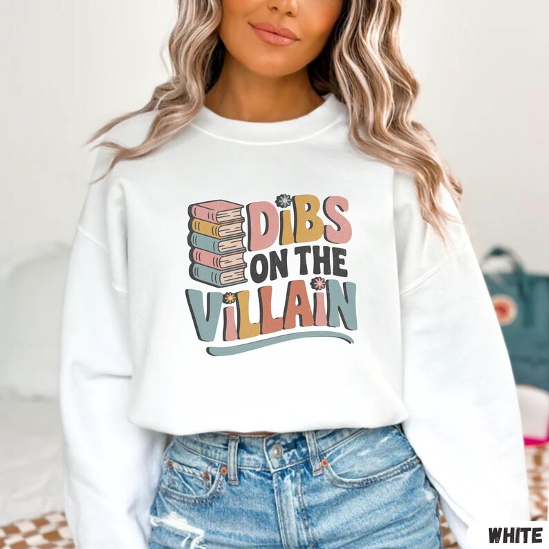 May include: White sweatshirt with the text "Dibs on the Villain" in a retro font. The design includes a stack of books and floral accents. The sweatshirt is a crew neck style.