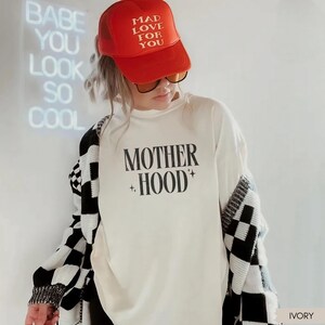 May include: A person wearing a red trucker hat with the text "MAD LOVE FOR YOU" and sunglasses. They are wearing a white long-sleeve shirt with the text "MOTHER HOOD" and a black and white checkered cardigan. A neon sign reads "BABE YOU LOOK SO COOL".