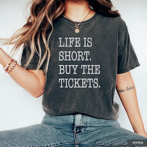 Puede incluir: Camiseta gris oscuro con el texto blanco "LIFE IS SHORT. BUY THE TICKETS.". La camiseta tiene un aspecto vintage y ligeramente descolorido. La modelo lleva vaqueros azules y un collar dorado.