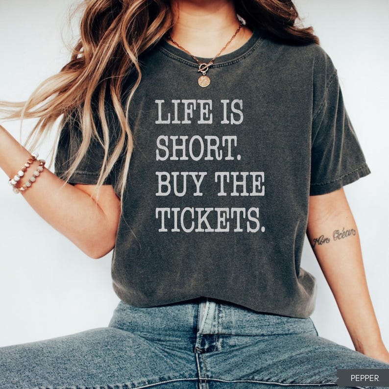 Puede incluir: Camiseta gris oscuro con el texto blanco "LIFE IS SHORT. BUY THE TICKETS.". La camiseta tiene un aspecto vintage y ligeramente descolorido. La modelo lleva vaqueros azules y un collar dorado.
