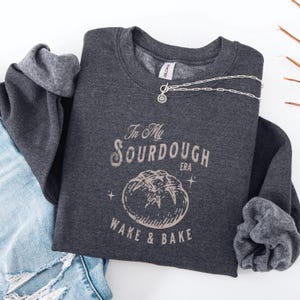 Peut inclure: Un sweat-shirt gris foncé avec le texte "In My Sourdough Era" et un graphique d'une miche de pain. Le texte "Wake & Bake" est sous le graphique du pain.