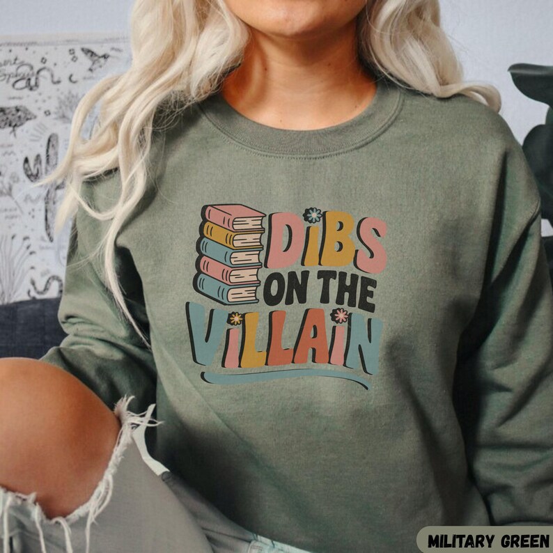 May include: Military green sweatshirt with the text "Dibs on the Villain" in a retro font, accompanied by a stack of colorful books. The sweatshirt is a solid color, and the text is in shades of pink, yellow, blue, and orange.