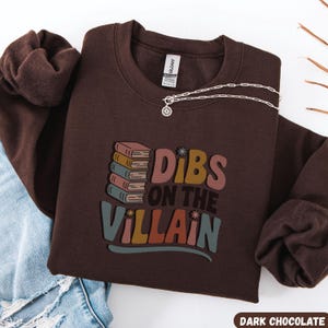 May include: A dark chocolate-colored sweatshirt with the text "Dibs on the Villain" in colorful retro lettering. The design includes a stack of books. A silver necklace with a pendant is draped on the sweatshirt.