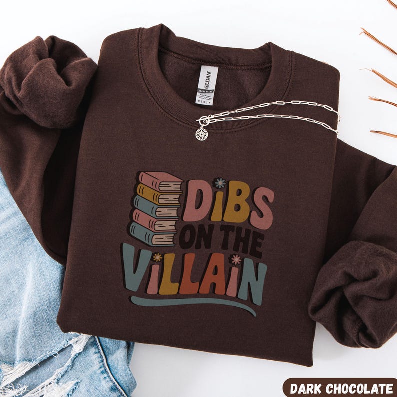 May include: A dark chocolate-colored sweatshirt with the text "Dibs on the Villain" in colorful retro lettering. The design includes a stack of books. A silver necklace with a pendant is draped on the sweatshirt.