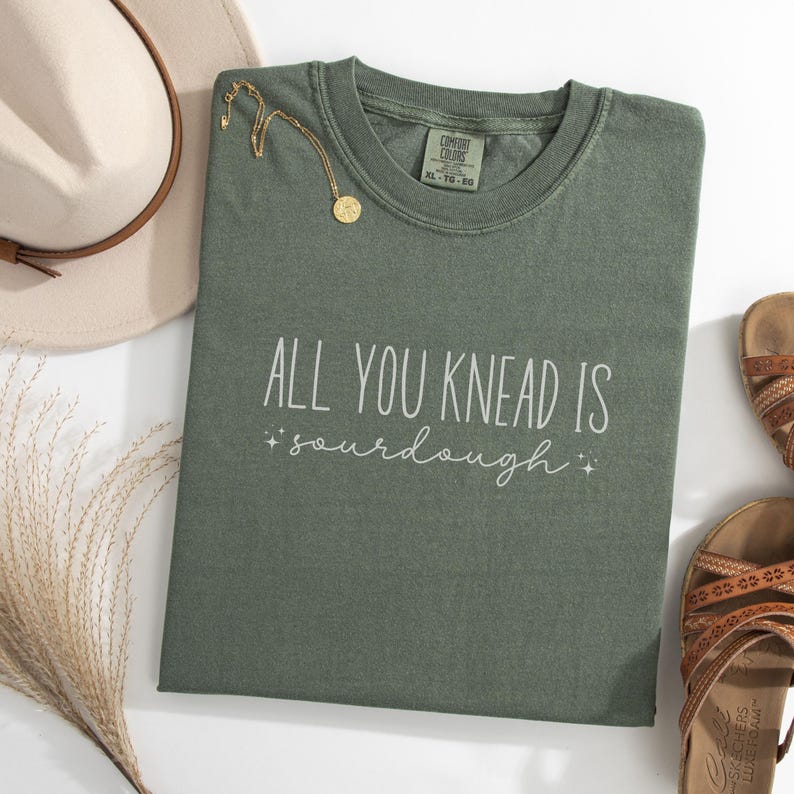May include: A green t-shirt with the text "All you knead is sourdough" in white lettering. The shirt has a distressed look and is laid flat on a white surface.