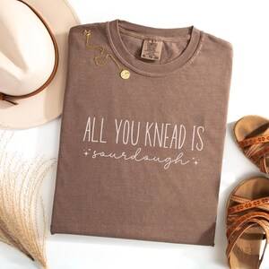 May include: A brown t-shirt with the text "All you knead is sourdough" in white lettering. The shirt has a distressed look and is made of a soft, comfortable fabric.