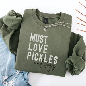 Peut inclure: Un sweat-shirt vert avec le texte "MUST LOVE PICKLES and dogs" imprimé sur le devant. Le texte est en police blanche vieillie.