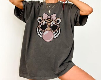 May include: Dark grey t-shirt featuring a cartoon tiger face with heart-shaped sunglasses, a pink bow, and a bubblegum bubble. The shirt has a relaxed fit and short sleeves. The word "PEPPER" is at the bottom.