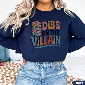 May include: Navy blue sweatshirt with the text "Dibs on the Villain" in a retro font. The design includes a stack of books in shades of pink, orange, and blue. A casual, comfortable top for book lovers.