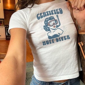 May include: White t-shirt with the text "CERTIFIED MUFF DIVER" in blue. The shirt features a retro illustration of a woman wearing a diving mask and snorkel. The shirt is a crew neck style.