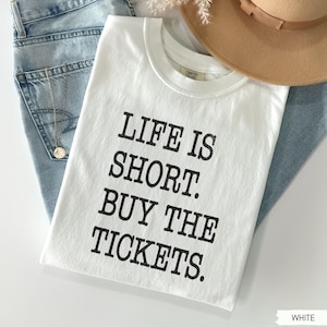 Puede incluir: Una camiseta blanca con el texto "LIFE IS SHORT. BUY THE TICKETS." en negro. La camiseta est&aacute; doblada y colocada sobre unos vaqueros azules, con un collar dorado y un sombrero marr&oacute;n en el fondo.
