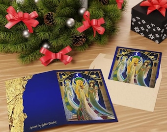 Puede incluir: Tarjetas de felicitación con una ilustración de ángeles, dentro de un sobre azul con detalles de pan de oro. La obra de arte está sobre una superficie de madera con una corona navideña, lazos rojos y una caja de regalo negra con un lazo rojo.