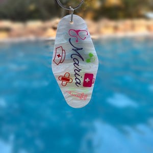 May include: A white and grey marbled acrylic motel-style keychain featuring the name Maria in cursive script, surrounded by medical-themed icons including a stethoscope, nurse cap, bandage, pill, first aid kit, and syringe in various colors.