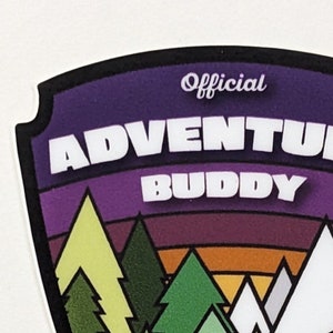 May include: A purple, yellow, green, blue, and white illustrated sticker with a black border. The sticker is shaped like a shield and features a mountain range, trees, and a river. The text "Official Adventure Buddy" is printed in white on the sticker.