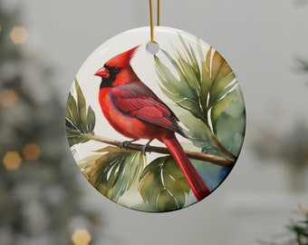 May include: Round ceramic Christmas ornament featuring a watercolor painting of a vibrant red cardinal perched on a branch with green leaves. The ornament has a gold string for hanging and is set against a blurred background.