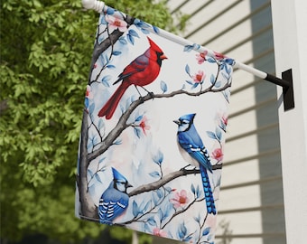 May include: A white garden flag with a watercolor design of a cardinal and two blue jays perched on branches with pink blossoms. The background is a soft blue and white watercolor wash.