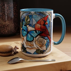 May include: A white ceramic mug with a blue rim and handle. The mug features a stained glass design of a cardinal, butterfly, and roses.