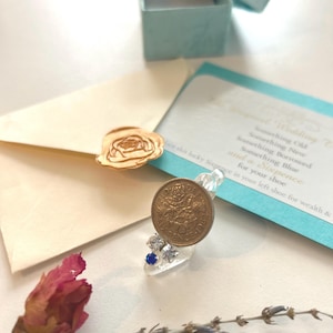 May include: A clear resin ring featuring an embedded vintage sixpence coin, a small blue rhinestone, and two clear rhinestones. The ring is displayed with a light blue gift box, a cream envelope with a gold rose seal, and a card with wedding text.