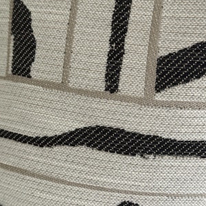 May include: Close-up of a textured fabric with a neutral beige background. The design features bold black and light brown stripes and curved shapes, creating a modern, abstract pattern. The fabric appears to be woven.
