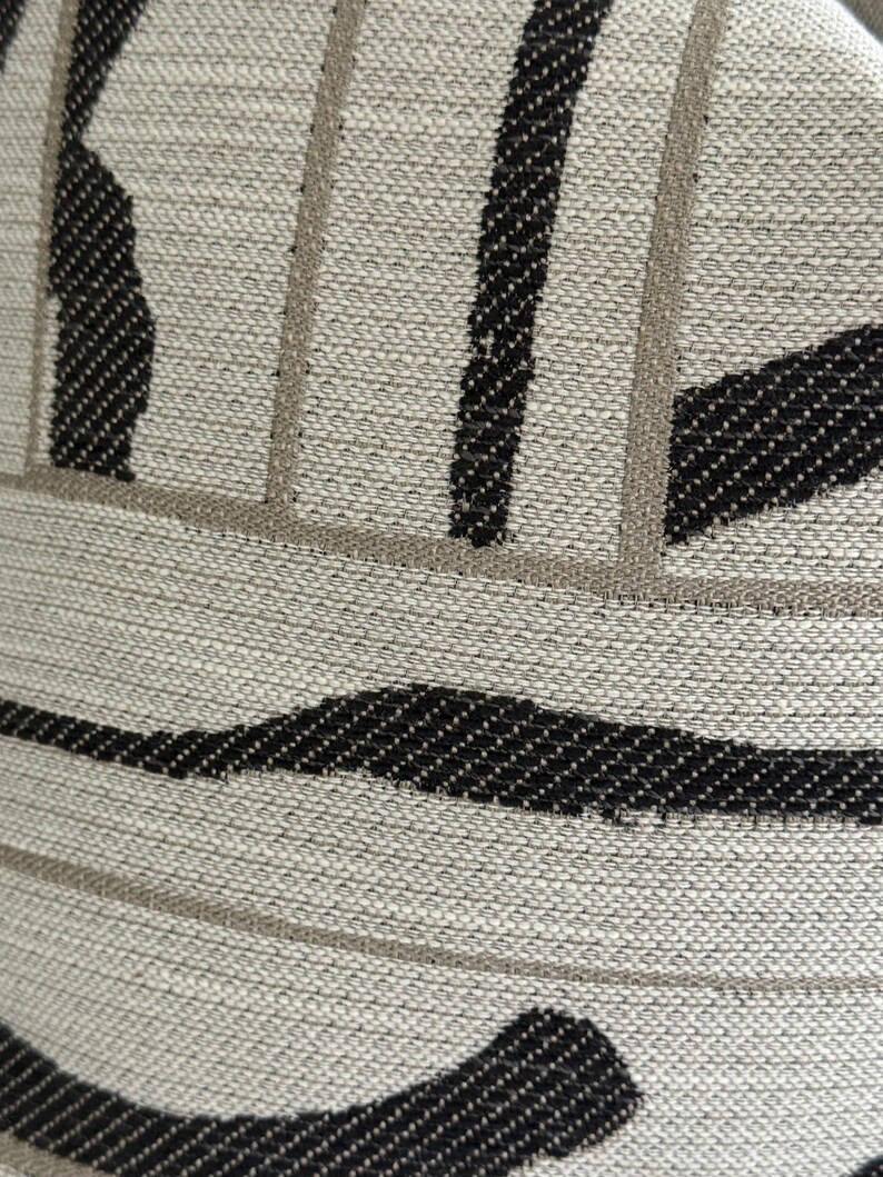 May include: Close-up of a textured fabric with a neutral beige background. The design features bold black and light brown stripes and curved shapes, creating a modern, abstract pattern. The fabric appears to be woven.