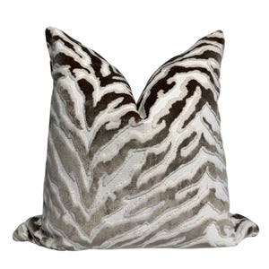 Puede incluir: Un cojín decorativo cuadrado con un estampado de rayas de cebra. Los colores principales son marrón, beige y blanco. El cojín tiene una textura suave y está diseñado para añadir un toque decorativo.