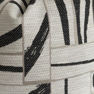 May include: Close-up of a decorative pillow with a textured, abstract design. The pillow features a neutral, light gray background with bold black brushstroke patterns and thin, light brown lines creating a geometric grid.