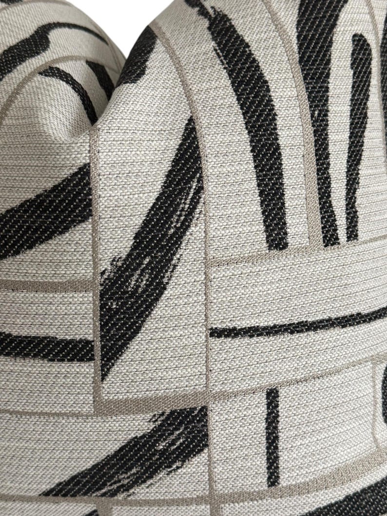 May include: Close-up of a decorative pillow with a textured, abstract design. The pillow features a neutral, light gray background with bold black brushstroke patterns and thin, light brown lines creating a geometric grid.