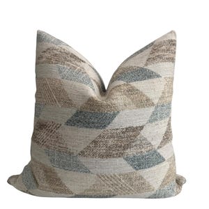 May include: A square throw pillow featuring a geometric pattern of interlocking diamonds and triangles. The fabric displays a textured weave in shades of beige, tan, light brown, and muted blue-grey, creating a repeating multi-dimensional design.