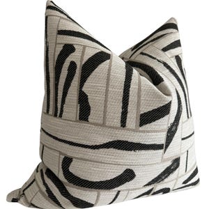 May include: Decorative throw pillow with a geometric pattern. The pillow features a cream-colored background with a repeating pattern of black and gray lines. The pillow is square and has a soft, textured appearance.