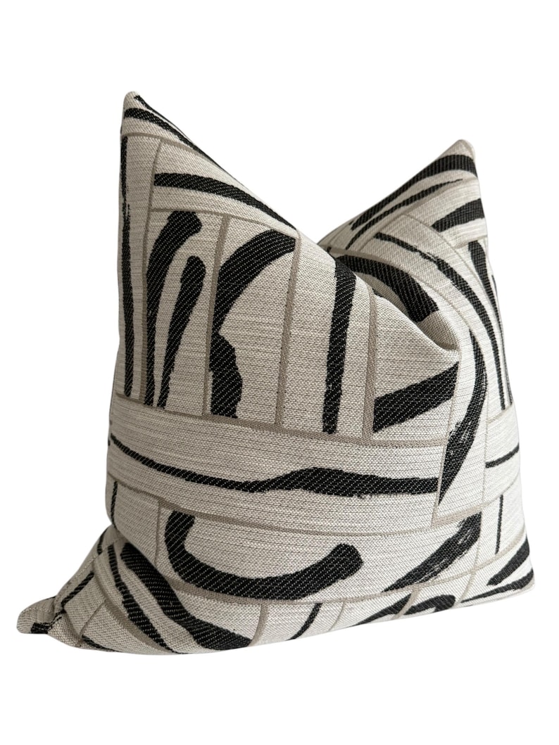May include: Decorative throw pillow with a geometric pattern. The pillow features a cream-colored background with a repeating pattern of black and gray lines. The pillow is square and has a soft, textured appearance.
