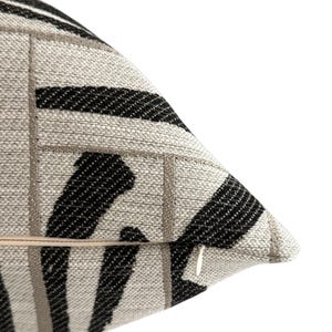 May include: Close-up of a decorative pillow with a textured fabric. The pillow features a pattern of black and gray stripes and squares on a light beige background. A zipper is visible on the side. The pillow is designed for home decor.