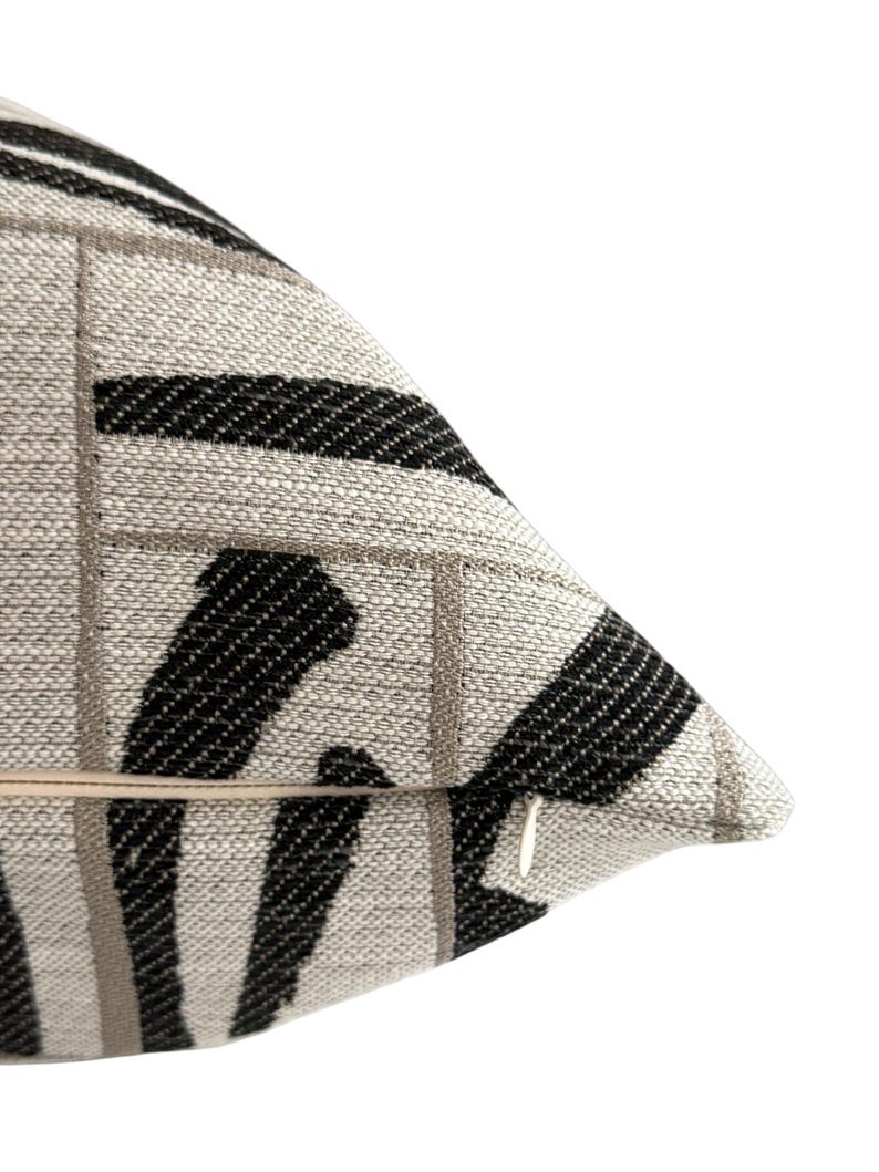 May include: Close-up of a decorative pillow with a textured fabric. The pillow features a pattern of black and gray stripes and squares on a light beige background. A zipper is visible on the side. The pillow is designed for home decor.