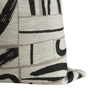 May include: Close-up of a decorative pillow with a geometric pattern. The pillow features a light beige background with black and gray brushstroke-style designs and lines. The fabric has a textured appearance, adding depth to the design.