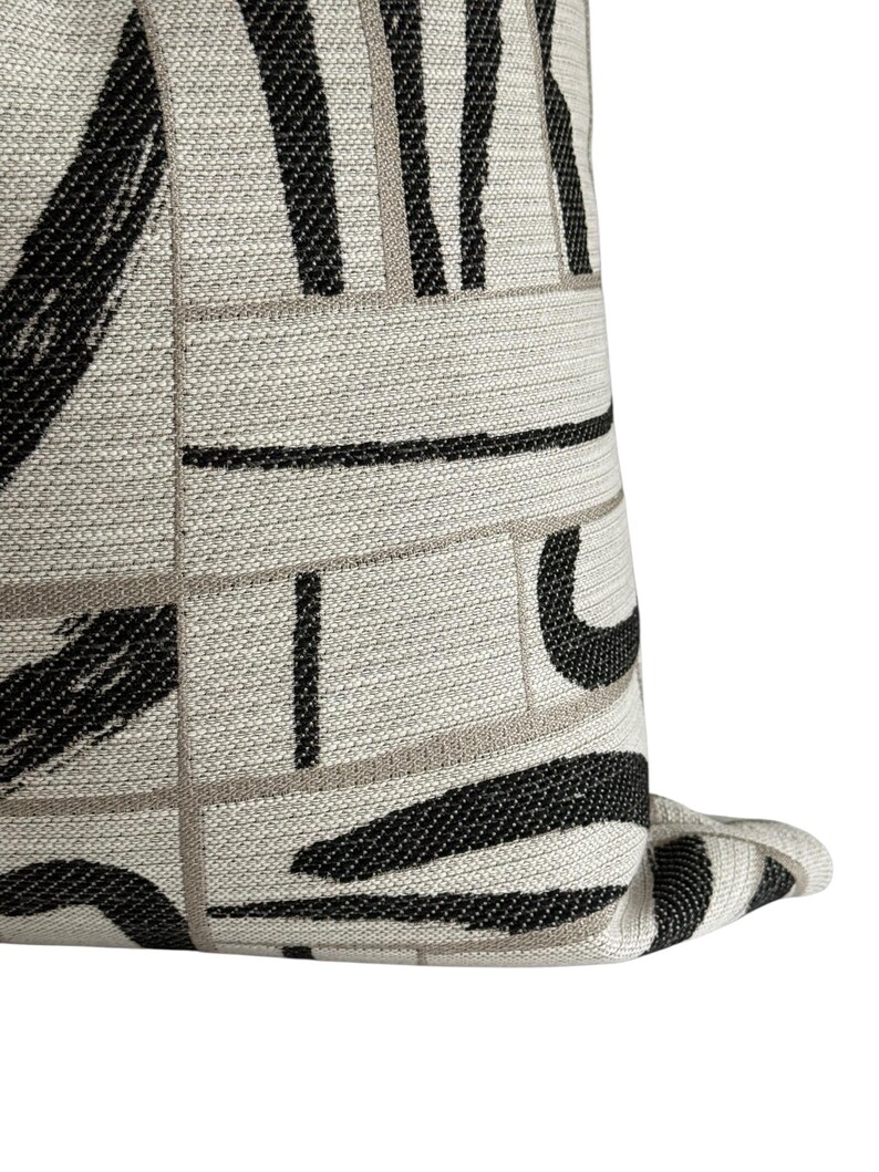 May include: Close-up of a decorative pillow with a geometric pattern. The pillow features a light beige background with black and gray brushstroke-style designs and lines. The fabric has a textured appearance, adding depth to the design.